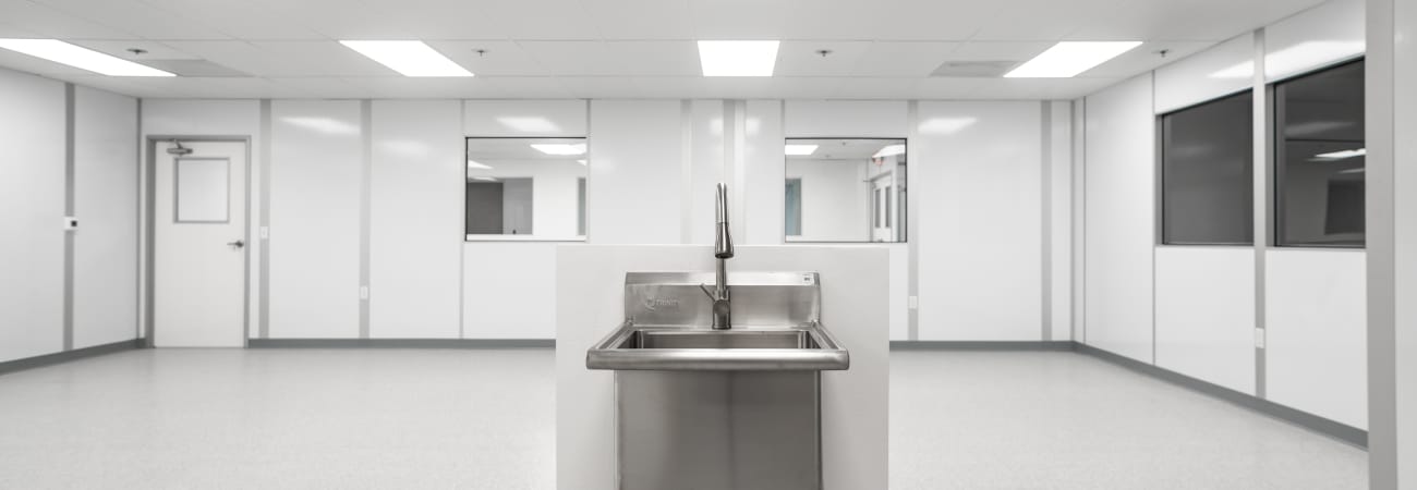 Stainless steel sink in the center of a bright, sterile cleanroom with white walls, large windows, and overhead fluorescent lighting.
