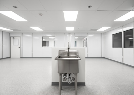 Stainless steel sink in the center of a bright, sterile cleanroom with white walls, large windows, and overhead fluorescent lighting.