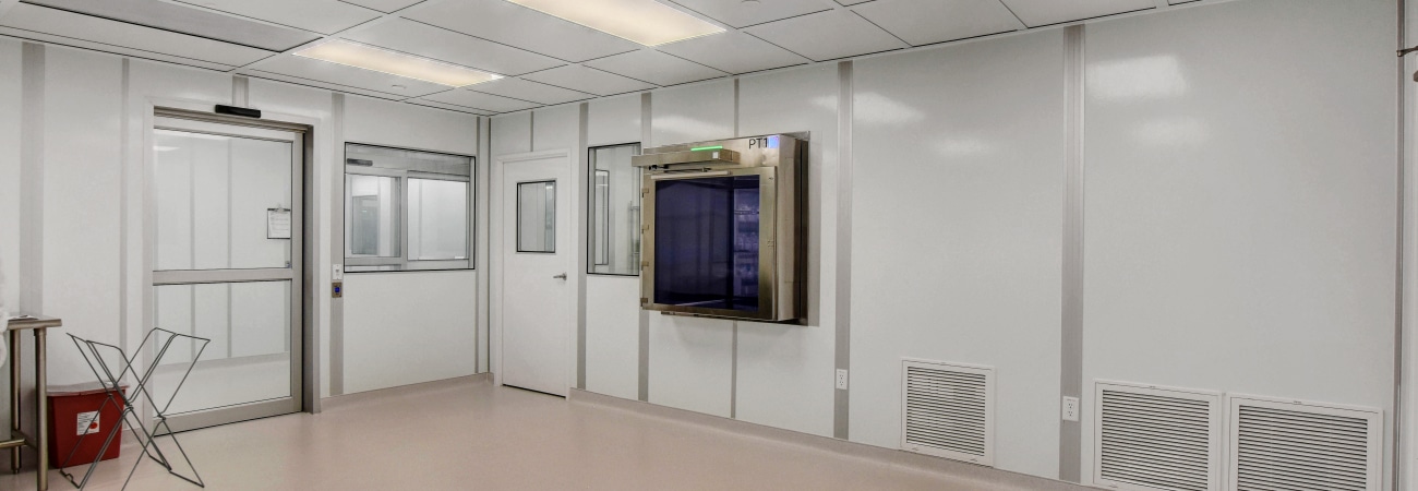 Modular cleanroom by Allied Cleanrooms with white panel walls, a smooth flooring system, and a grid ceiling with integrated lighting. The room features a pass-through chamber, multiple doors, and ventilation panels, designed for controlled environments requiring high cleanliness standards.