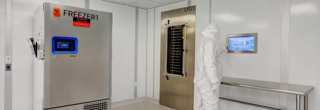 Modular cleanroom by Allied Cleanrooms featuring a technician in full gowning operating a touchscreen interface. The space includes a stainless steel lyophilizer unit labeled "LY01" and an ultra-low temperature freezer labeled "FREEZER1" set to -79°C, all housed in a clean, controlled environment with integrated lighting and seamless finishes.