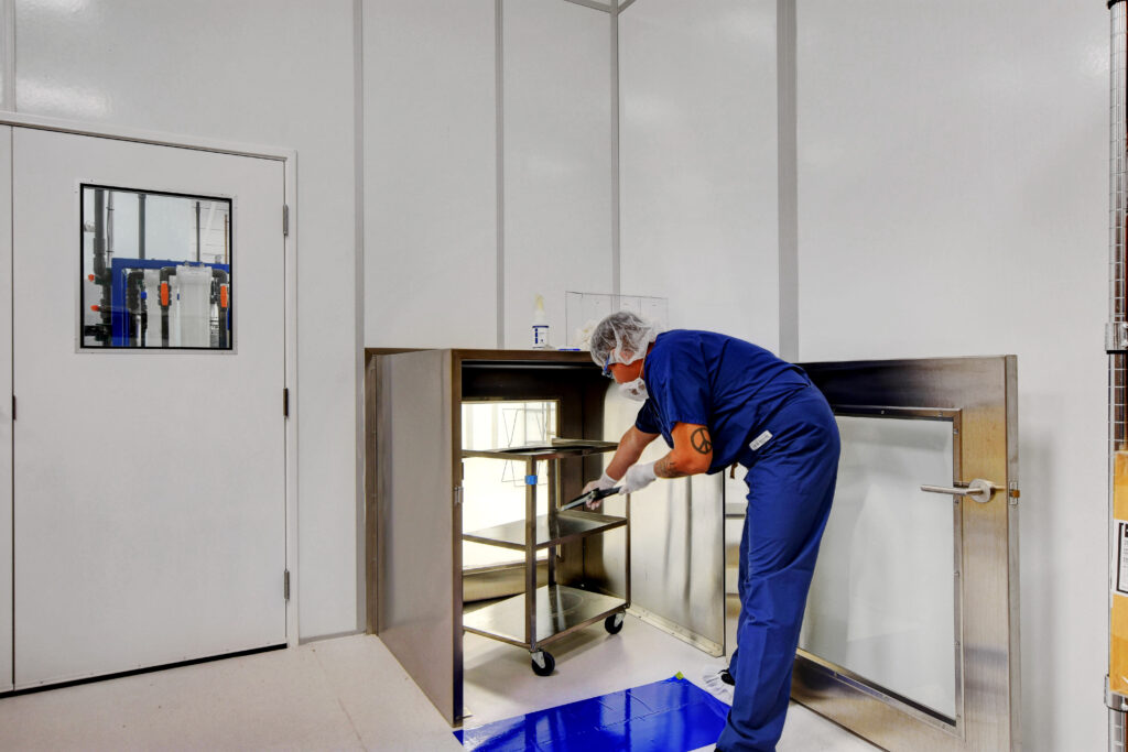 A cleanroom technician in protective garments is using a pass-through chamber to maintain sterile conditions.
