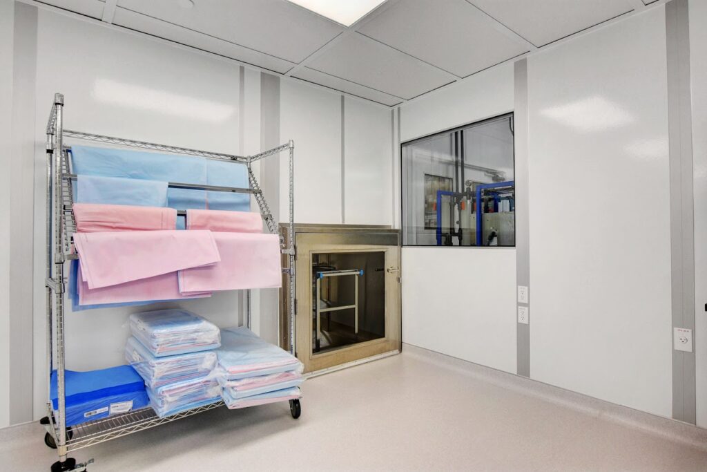 Sterile cleanroom environment showing a metal shelving unit with neatly stacked blue and pink fabrics, adjacent to a glass window revealing industrial equipment.