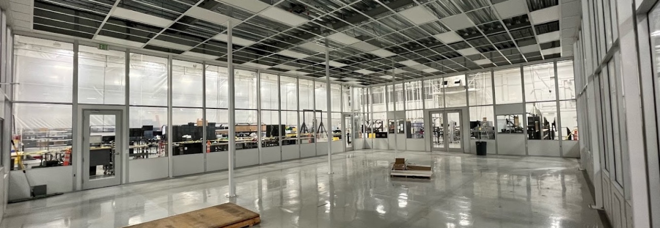 In-progress modular cleanroom by Allied Cleanrooms featuring glass wall panels, a partially installed ceiling grid, and a high-gloss epoxy floor. The cleanroom overlooks an active manufacturing area and is designed for scalable, controlled environments.