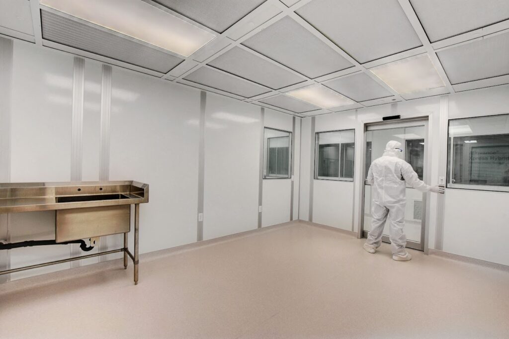 A person in a full-body white cleanroom suit stands facing the door in a sterile cleanroom with a stainless steel sink and under bright overhead lights.
