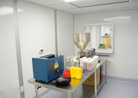 A cleanroom with sterile white walls, a blue machine, a large metal hopper, and various containers on a metal table, adhering to cGMP or GMP standards for pharmaceutical or chemical manufacturing.