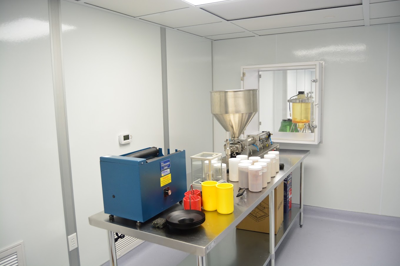 A cleanroom with sterile white walls, a blue machine, a large metal hopper, and various containers on a metal table, adhering to cGMP or GMP standards for pharmaceutical or chemical manufacturing.