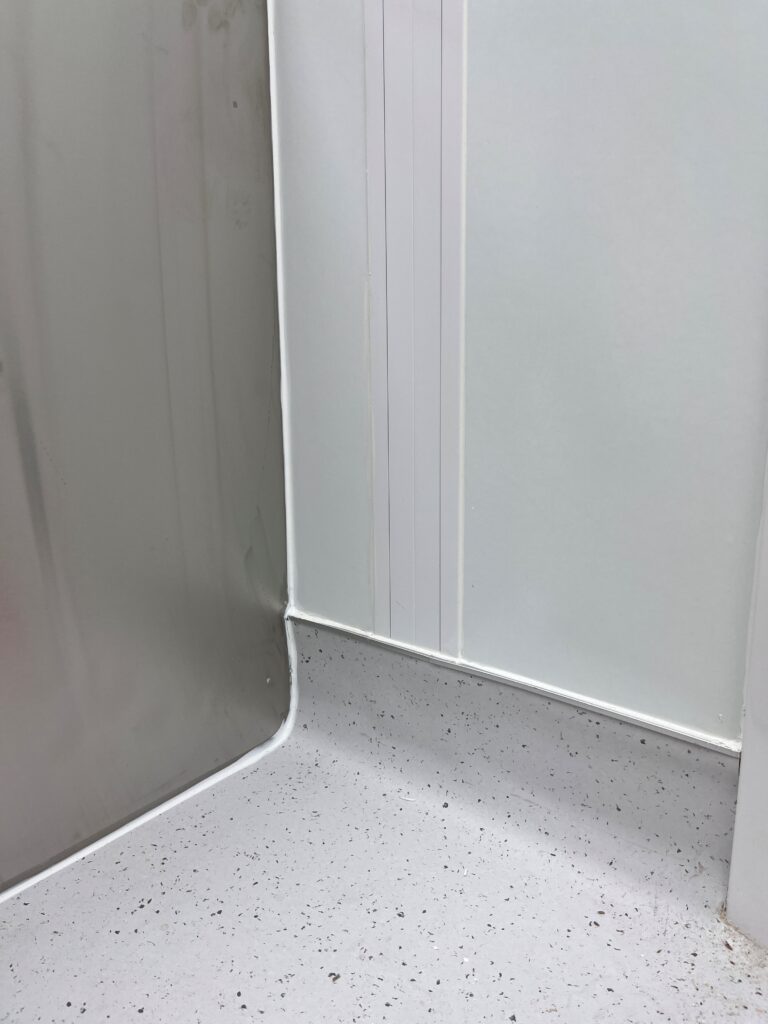 Cleanroom corner with seamless coving, smooth wall panels, and speckled flooring.