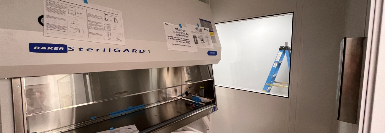 A laminar flow hood inside a cleanroom, labeled SterilGARD, with instructions posted on the front panel and a clear viewing window on the right wall. The workspace includes a reflective stainless steel surface, with a cardboard sheet on the floor in the foreground.