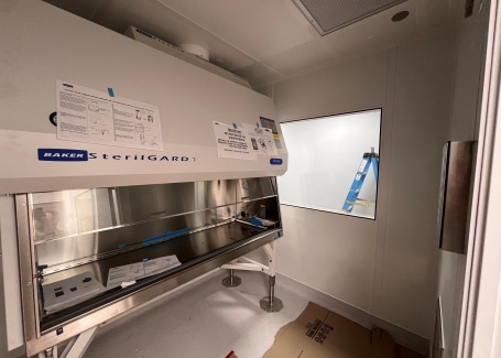 A laminar flow hood inside a cleanroom, labeled SterilGARD, with instructions posted on the front panel and a clear viewing window on the right wall. The workspace includes a reflective stainless steel surface, with a cardboard sheet on the floor in the foreground.