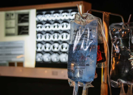 An image of intravenous (IV) bags, one containing a blue liquid and another with a red liquid, in a hospital setting with medical monitors displaying patient data in the background.